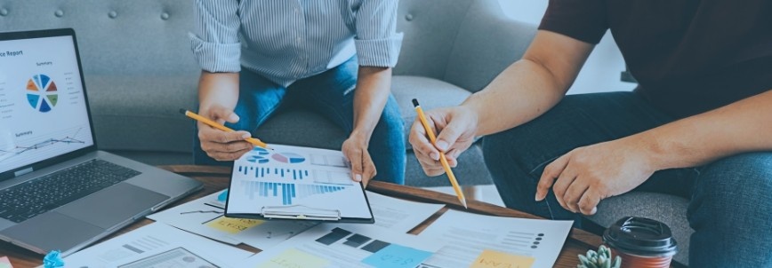 Two people sit at a table covered with charts, graphs, and papers, reviewing data and taking notes. Nearby, a laptop displays Financial Forecasting with BVDash, alongside a coffee cup and sticky notes.