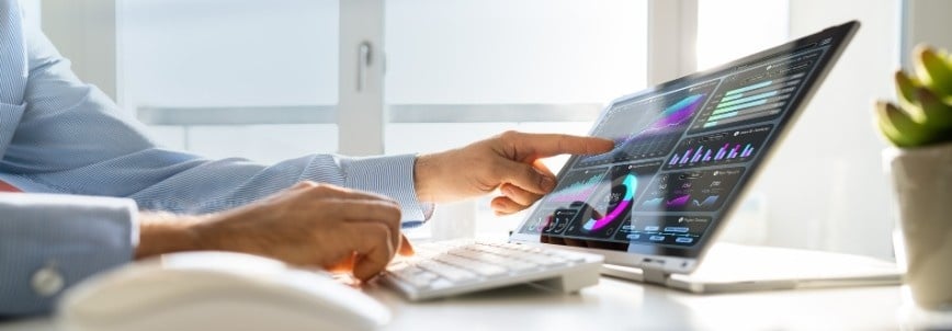 A person points at colorful data visualizations and charts displayed on a laptop screen, enhancing client engagement with BVDash, while another hand types on a keyboard in a bright office filled with natural light.