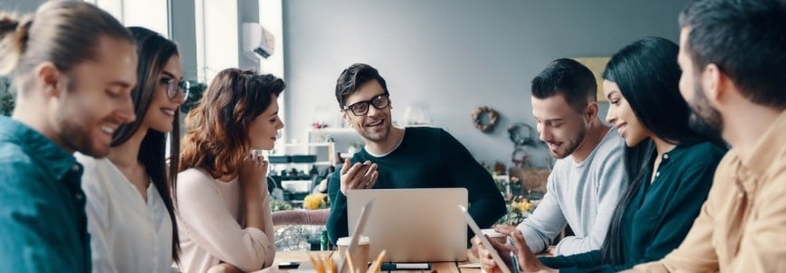 A group of seven young adults sit around a table in a modern office, smiling and talking. Laptops, notebooks, and coffee cups are on the table, hinting at a collaborative sprint planning or brainstorming session.
