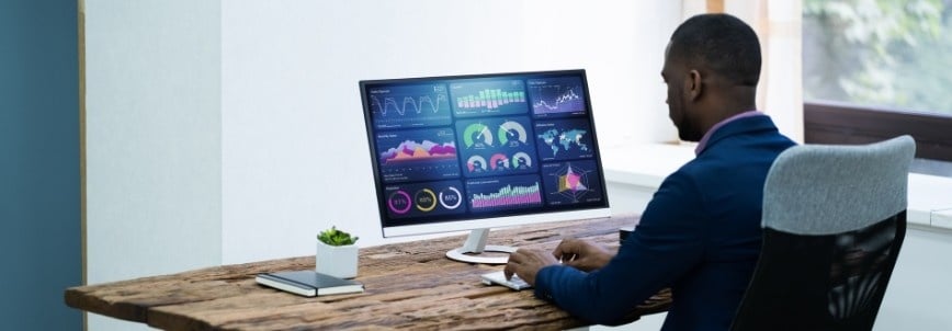 A man in a business suit sits at a wooden desk, analyzing colorful graphs and charts displayed on a large computer monitor as he manages Recurring Tasks in Notion in a modern office setting.