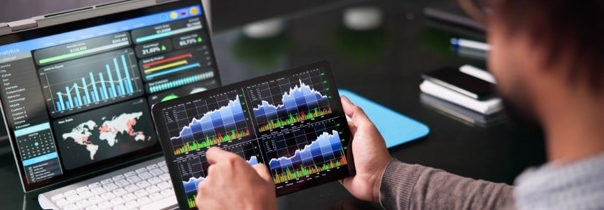A person looks at financial charts and graphs on a tablet, with more data and analytics displayed on a laptop in the background, suggesting financial analysis or trading work organized with Notion Pre-Built Templates.