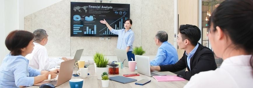 A woman presents financial analysis data on a large screen to five colleagues seated around a conference table with laptops, notepads, and plants in a modern office, highlighting insights aligned with key PMP domains.