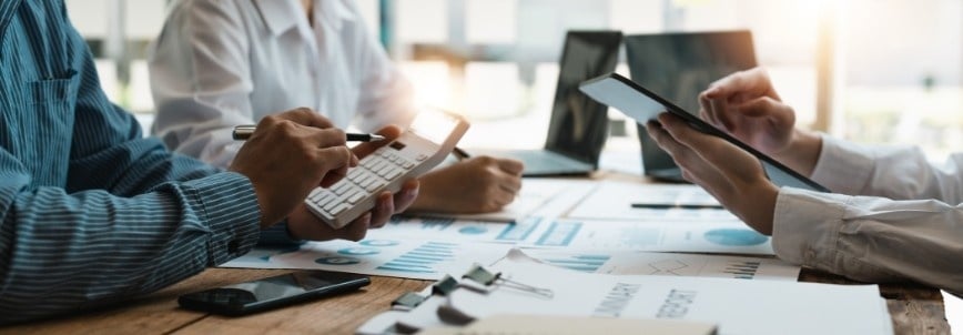 Three people sit at a table working with documents, charts, a calculator, and digital tablets. Sunlight streams in as they discuss and analyze financial data and the Project Budget in BVDash in an office setting.