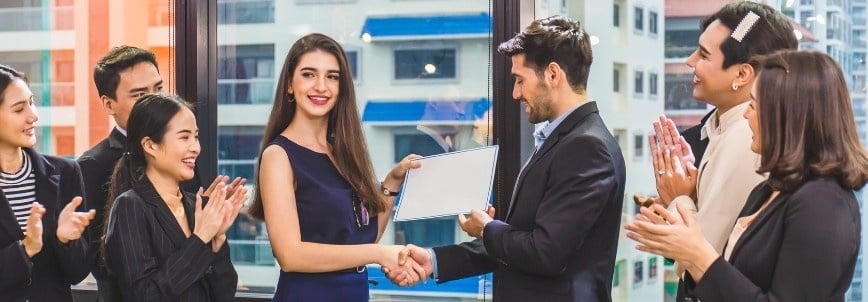 A woman receives a PMI Certification certificate from a man while shaking hands. Four colleagues stand around them, clapping and smiling in a modern office with large windows and city buildings in the background.