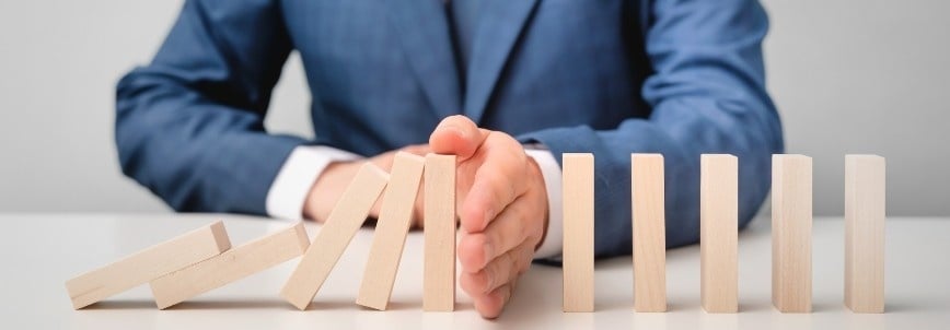 A person in a blue suit stops a row of falling wooden dominoes with their hand, symbolizing effective risk management in Agile vs Waterfall by preventing the remaining standing dominoes from toppling.