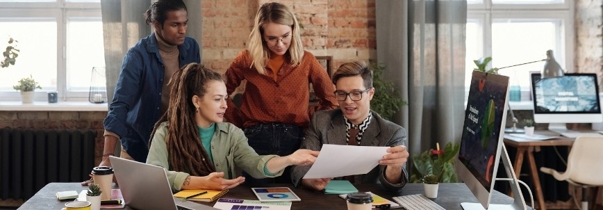 Four people are gathered around a desk in an office, discussing a document and collaborating on a project. Their teamwork highlights an Agile approach focused on the point of value delivery, with a computer and papers spread out before them.