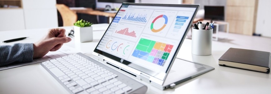 A person works at a desk with a white keyboard and an open laptop displaying colorful charts and graphs, using Notion Progress Bars for tracking data analysis. A plant, closed notebook, and pen are also on the desk.