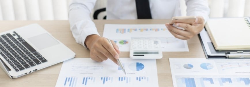 A person in business attire sits at a desk with charts, a laptop, PMP Sample Questions, a calculator, and documents—analyzing financial graphs while holding a smartphone and pointing at a report.