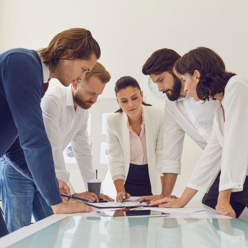 Five people in business attire stand around a table, closely looking at documents and discussing, suggesting a collaborative Sprint Planning meeting or team discussion in a modern office setting.