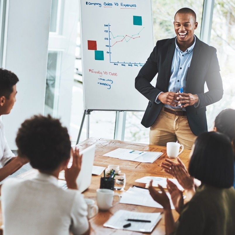 A man in business attire stands and smiles while giving a presentation to four colleagues seated at a conference table. Behind him, a flip chart displays a sales versus expenses graph, scribbled notes, and highlights key PMP domains.