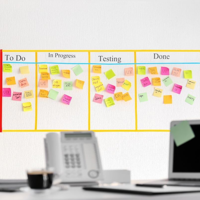 A Kanban board for Agile Planning features four columns—"To Do," "In Progress," "Testing," and "Done"—filled with colorful sticky notes. In the foreground, an office desk holds a phone, coffee cup, and scattered papers.