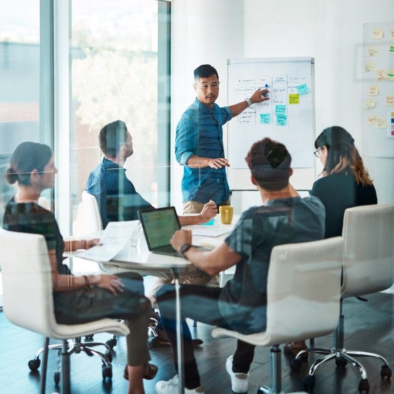 A man stands at a whiteboard giving a presentation on PMP domains to four colleagues seated around a conference table with laptops and papers in a modern office meeting room.