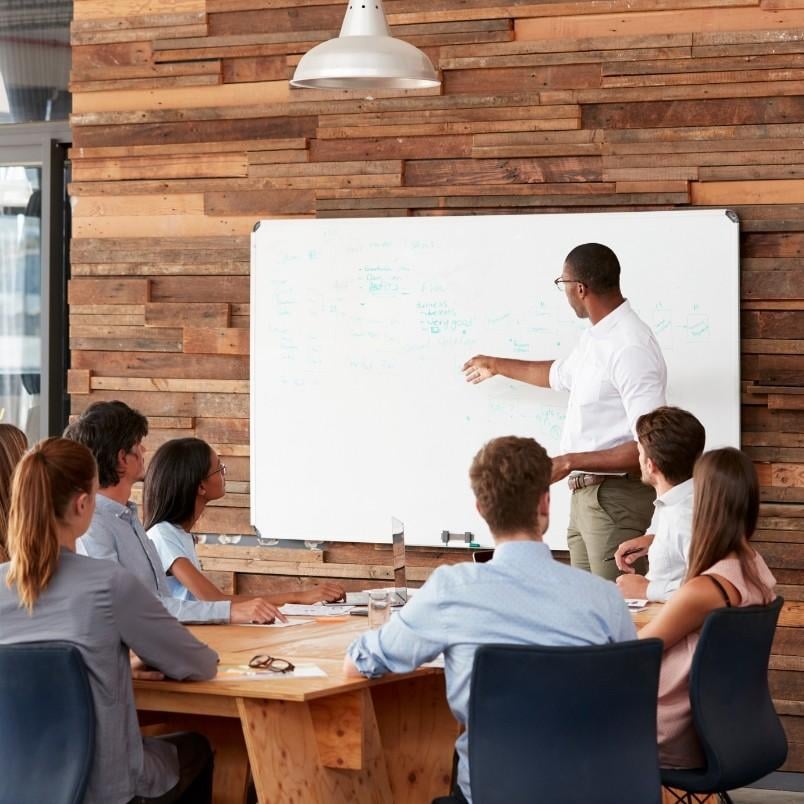 A man stands at a whiteboard, presenting to six colleagues in a modern office with wooden walls. The group listens attentively as he discusses strategies for stakeholder support, jotting key points on the board.