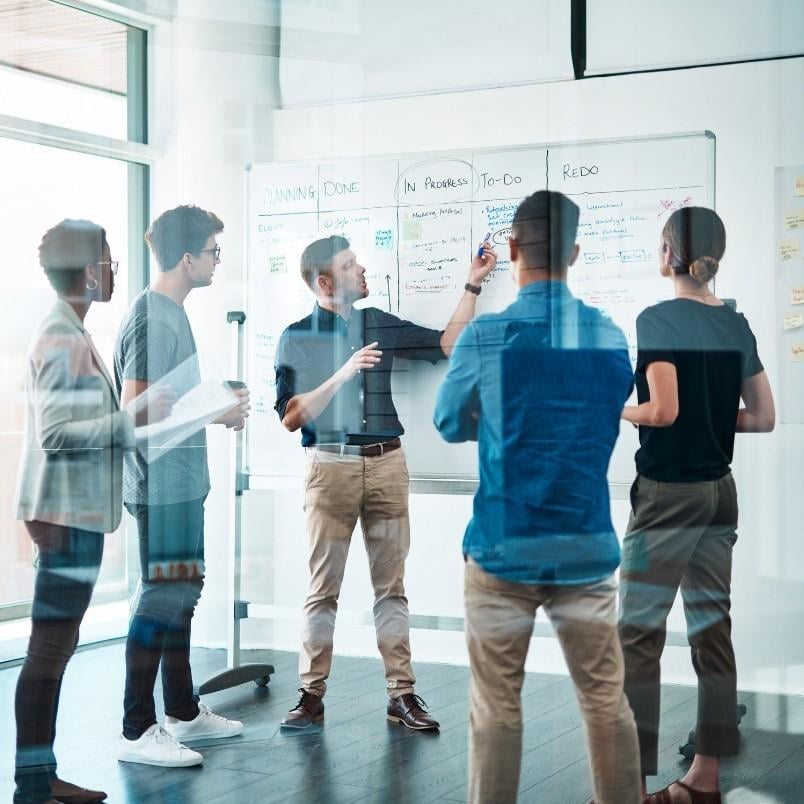 Five people stand and discuss around a whiteboard covered with notes and diagrams in a modern office, collaborating on a project; their reflections are visible in the glass walls as they reference their Company Wiki in Notion.