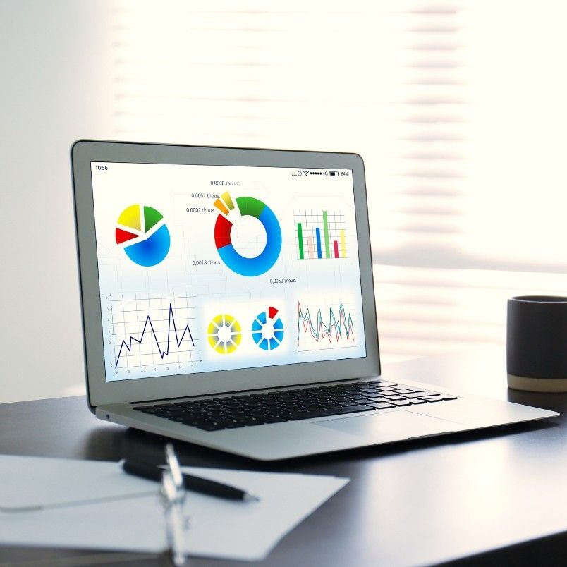 A laptop on a desk displays colorful charts, graphs, and Notion Progress Bars on its screen. Nearby are a pen, papers, and a cup, with sunlight streaming through window blinds in the background.