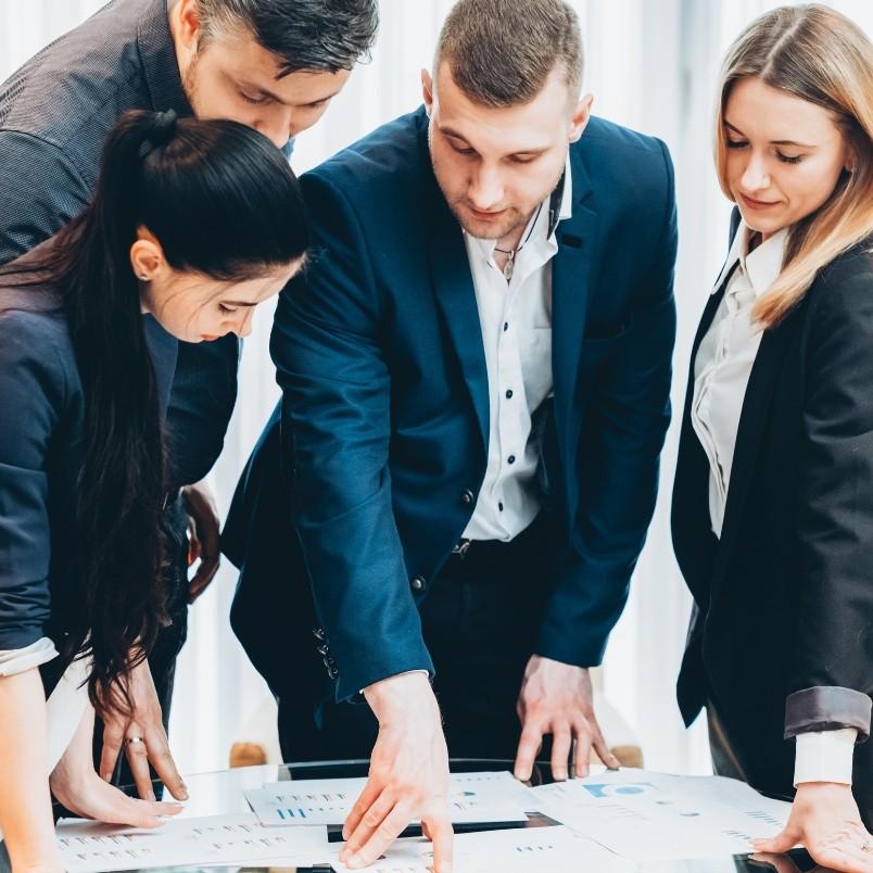Four business professionals in formal attire stand around a table, closely examining and discussing documents and charts related to PMP domains laid out in front of them.