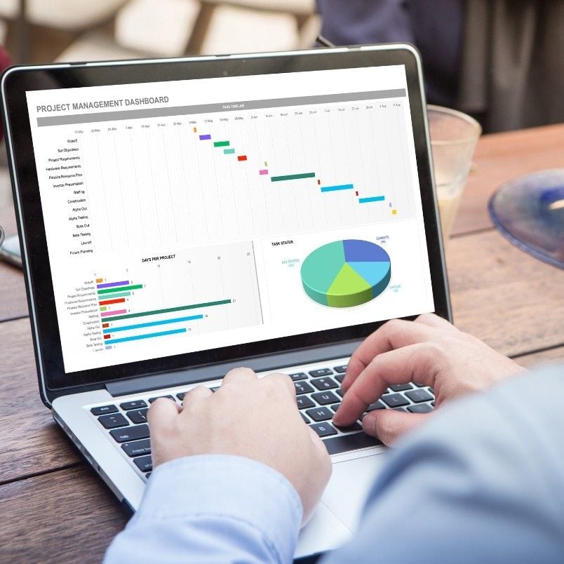 A person uses a laptop displaying a project management dashboard with a Gantt chart, bar graph, and pie chart for managing files in BVDash, while sitting at a wooden table.