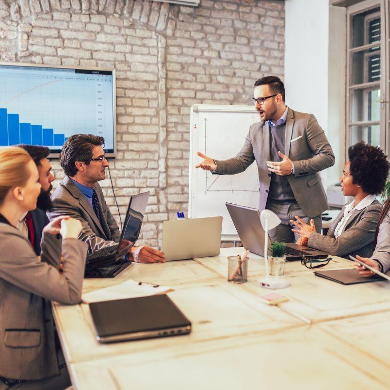 Five professionals in business attire have a meeting in a modern office, discussing Agile Planning. One man stands and gestures while speaking to four seated colleagues, with laptops, papers, charts on the table, and a graph displayed on a screen.