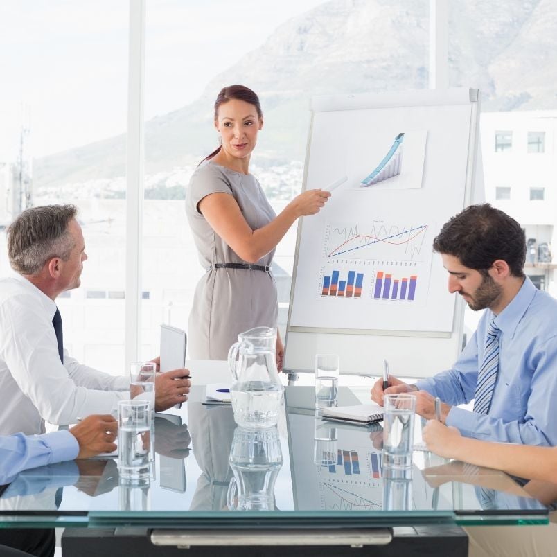 A woman stands by a flip chart with graphs, presenting Agile Planning strategies to three seated colleagues in a modern office with large windows and a mountain view. They appear focused and engaged in a business meeting.