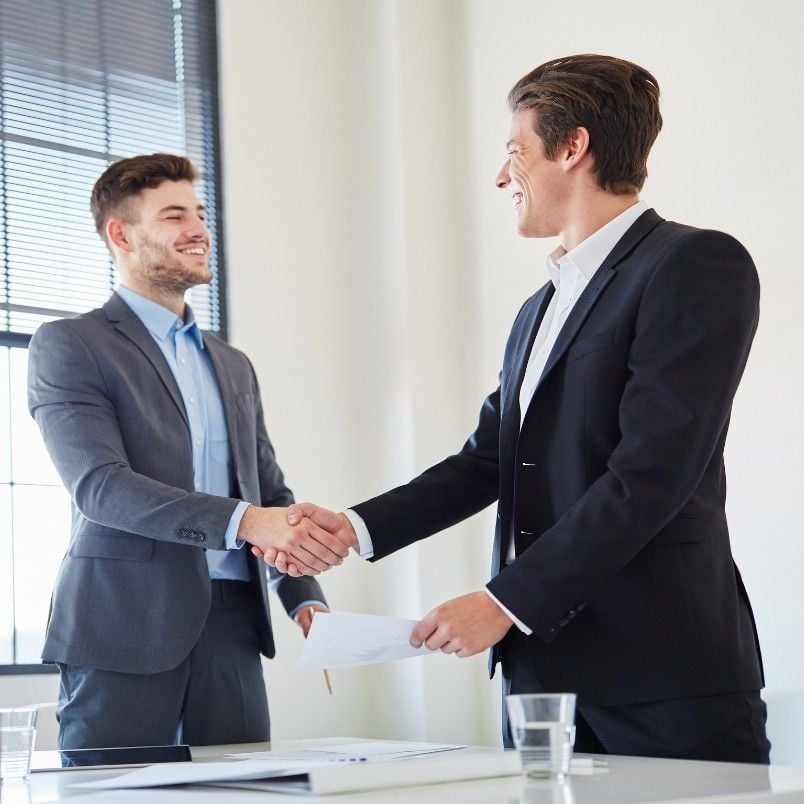 Two men in business suits smiling and shaking hands across a table, showing strong stakeholder support in a bright office setting with documents and glasses of water on the table.