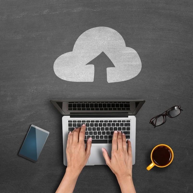 A person typing on a laptop with a cloud upload icon drawn on a chalkboard background, illustrating Managing Files in BVDash, with a smartphone to the left and a cup of coffee to the right.
