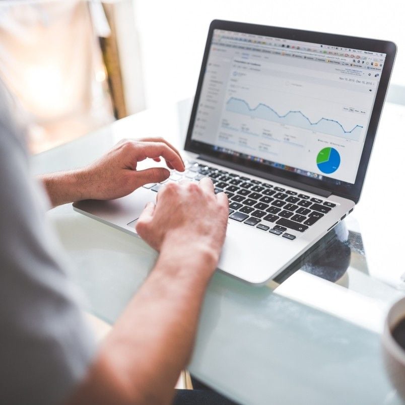 A person types on a laptop displaying analytics data, including graphs and charts on screen—possibly while managing files in BVDash. The laptop rests on a glass table, with a coffee cup partially visible in the corner.