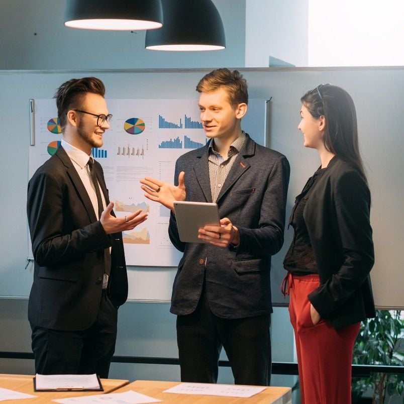 Three young professionals in business attire discuss the preparation of project details in an office. One holds a tablet while charts with graphs on a whiteboard highlight their planning process. They are smiling and engaged in conversation.