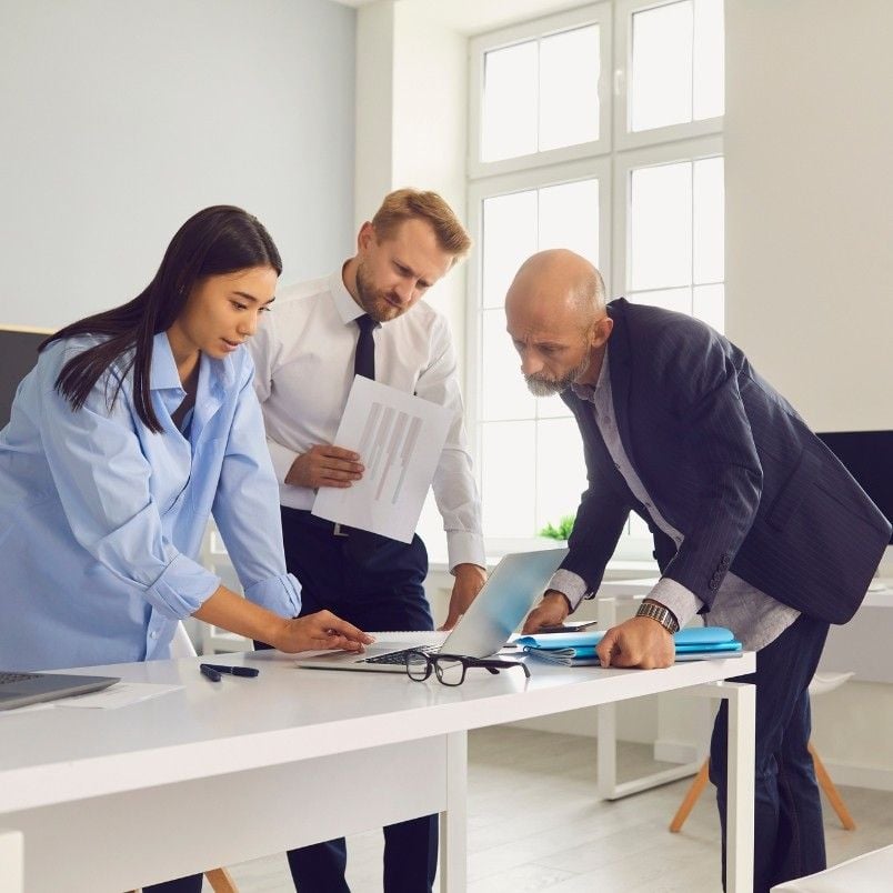 Three business people—two men and a woman—stand around a desk in an office, collaborating and looking at a laptop screen, discussing updates to their Company Wiki in Notion with documents and eyeglasses on the table.