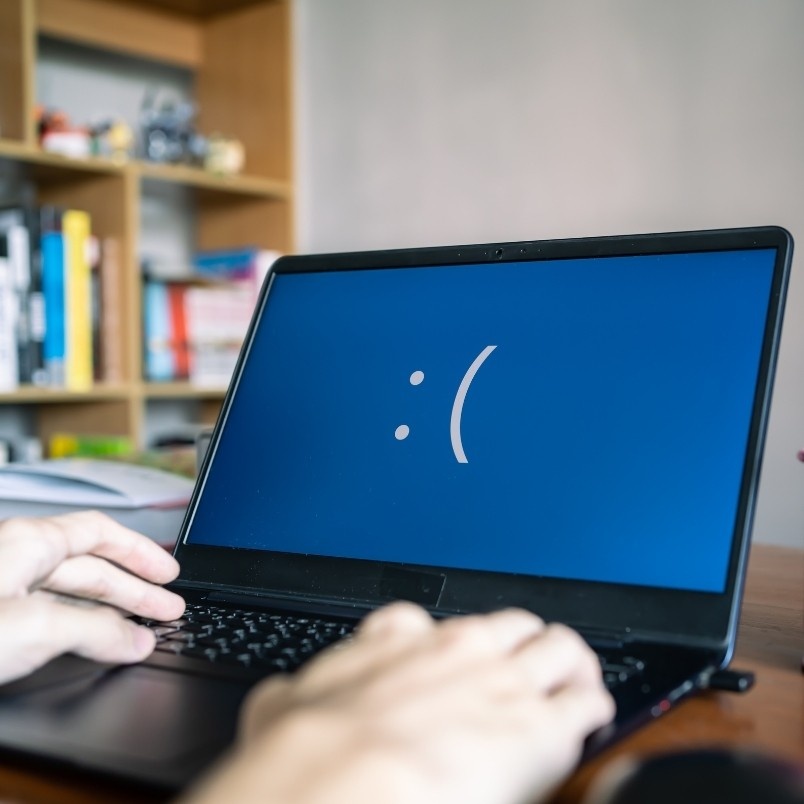 A person types on a laptop displaying a blue screen with a large white sad face emoticon, suggesting a system error. A bookshelf with books and items is in the background, perhaps hinting at researching the Pros and Cons of Onsite PMP Testing.