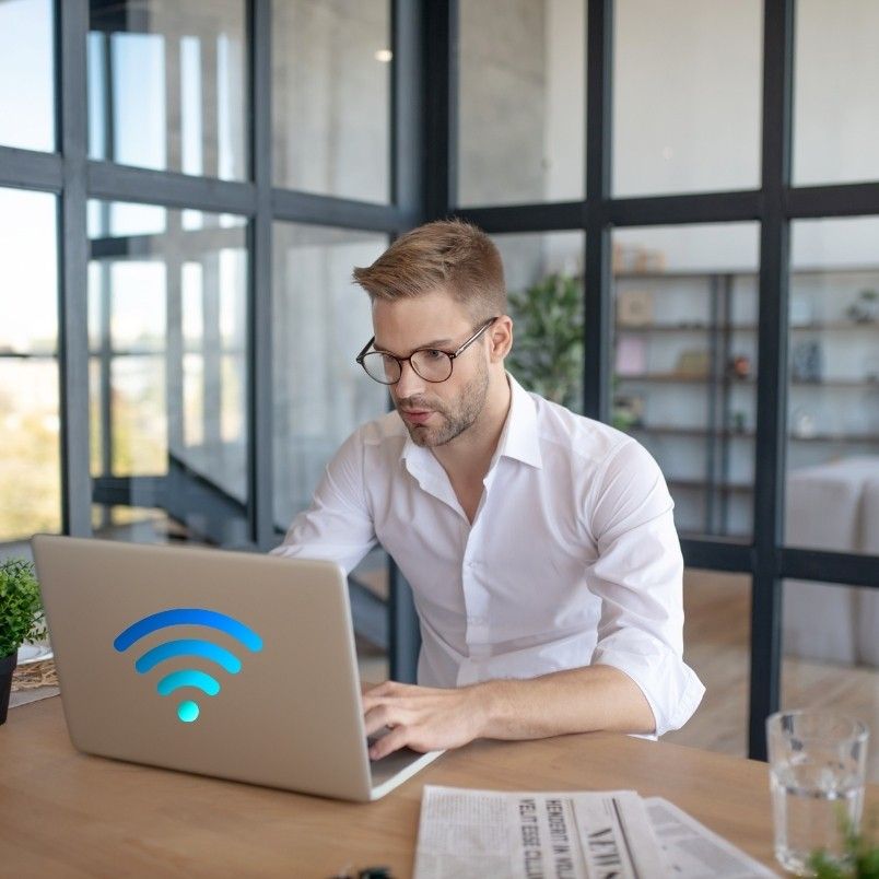 A man wearing glasses and a white shirt works on a laptop with a large blue Wi-Fi symbol, possibly researching the pros and cons of onsite PMP testing, seated at a desk in a modern, glass-walled office with papers and a glass of water nearby.