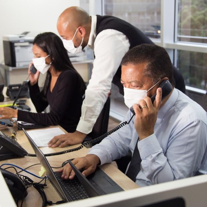 Three people wearing face masks work in an office; two are on the phone at their desks with laptops, while the third stands behind them looking at a document, perhaps discussing the pros and cons of onsite PMP testing.
