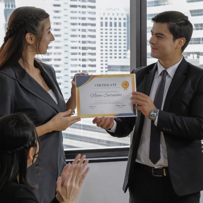 A man in a suit hands a certificate to a woman in business attire while another woman sits nearby clapping. They are in an office with large windows and city buildings, celebrating her achievement after learning about the CAPM® Certification Cost.