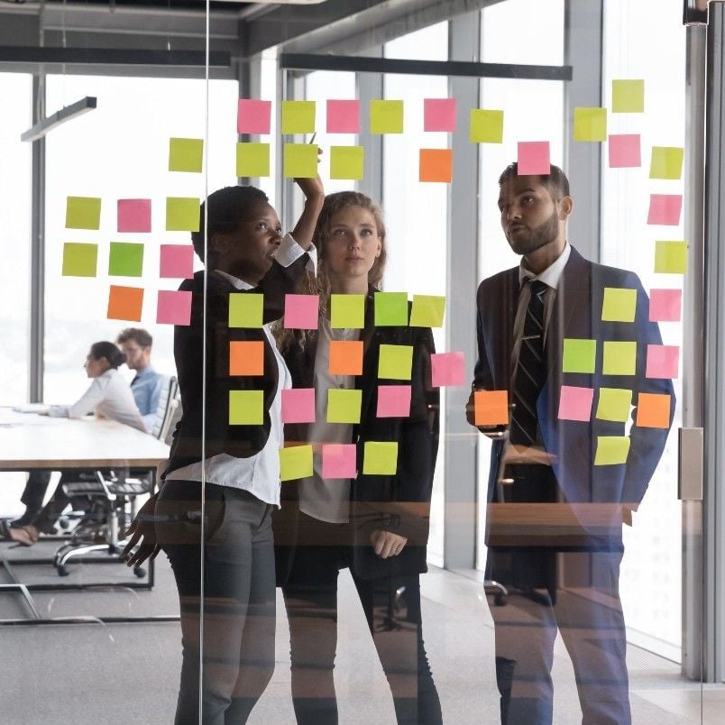 Three professionals stand in front of a glass wall covered with colorful sticky notes, brainstorming Types of Agile Projects in a modern office setting. Other colleagues are working at desks in the background.