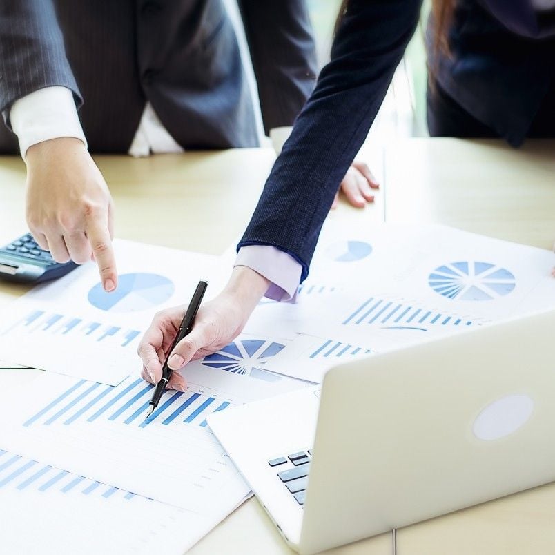 Two people in business attire review graphs and charts on paper at a desk with a laptop, one pointing and the other using a pen. Blue bar and pie charts are visible, suggesting financial analysis or a Project Charter meeting.