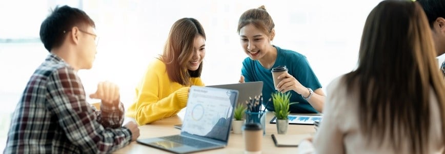 A group of young adults sit around a table with laptops, notebooks, and coffee, collaborating and smiling in a bright, modern workspace as they apply PMP principles to their project discussions.