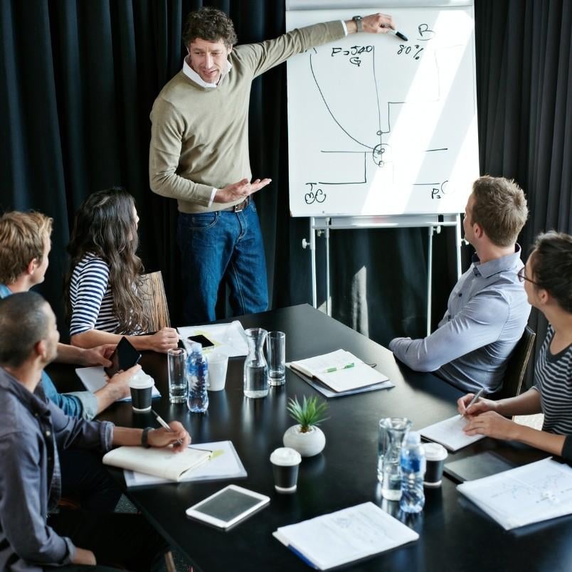 A Project Manager in Agile stands beside a flip chart with diagrams, presenting to five seated colleagues around a conference table with notebooks, coffee cups, water bottles, and papers.