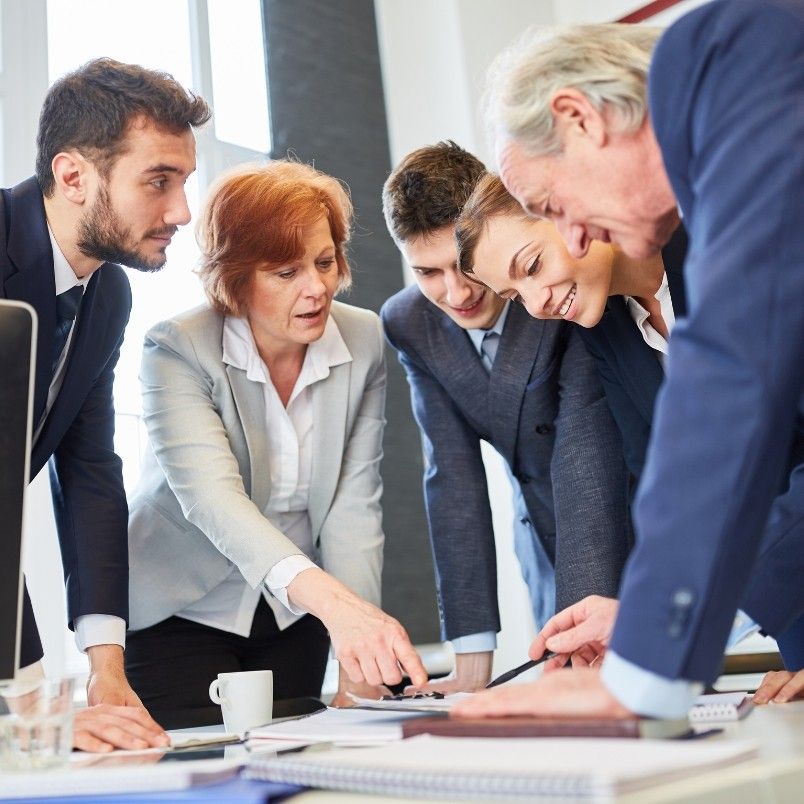 Five business professionals in suits gather around a desk, discussing documents and pointing at papers, appearing engaged as they review the types of Agile projects in a bright office setting.
