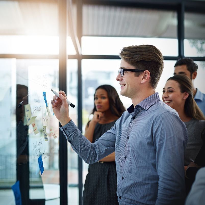 A man in glasses writes "What is Development Approach" on a transparent board with a marker, while colleagues stand nearby smiling and watching in a modern office setting.