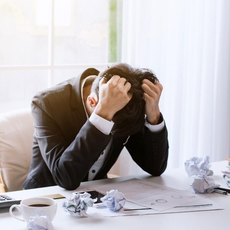 A person in a suit sits at a desk, head down and gripping their hair in frustration. Crumpled papers, documents labeled "CAPM Exam Preparation," a coffee cup, and a laptop are scattered on the desk, showing stress from studying.