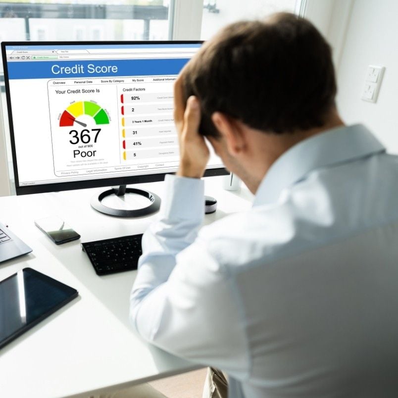 A man sits at a desk, holding his head in frustration while looking at a computer screen displaying a credit score of 367, labeled "Poor," alongside CAPM Exam Preparation materials and a colored gauge with credit factors.