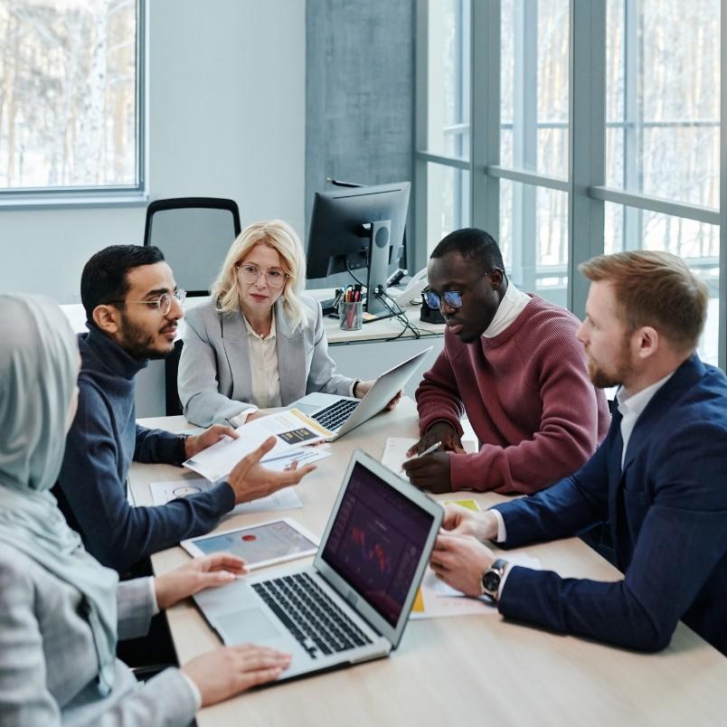 Five people of diverse backgrounds are sitting around a conference table with laptops and documents, discussing Types of Agile Projects in a business meeting held in a modern office with large windows.