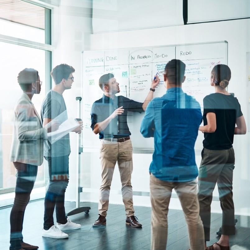 Five people, including a Project Manager in Agile, stand in front of a whiteboard covered with notes and diagrams, actively discussing and brainstorming ideas in a modern office with large windows.
