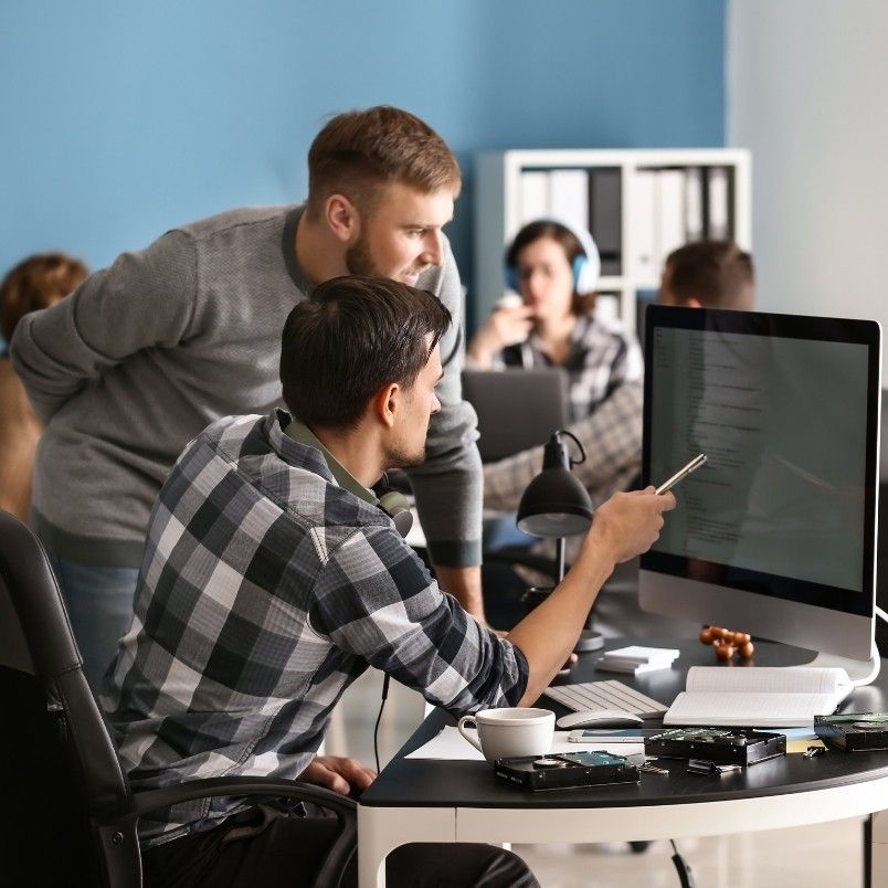 Two men are working together at a computer desk in an office, discussing the pros and cons of onsite PMP testing. One is seated and pointing at the monitor with a pen while the other stands, both focused on the screen. Other coworkers are visible in the background.