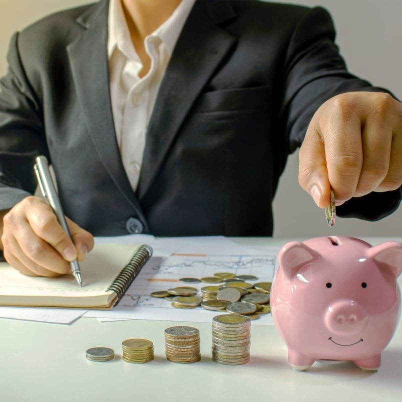 A person in a suit writes in a notebook, with coins, graphs, and a pink piggy bank in front. One hand drops a coin into the piggy bank, symbolizing saving or budgeting for the CAPM® Certification Cost.