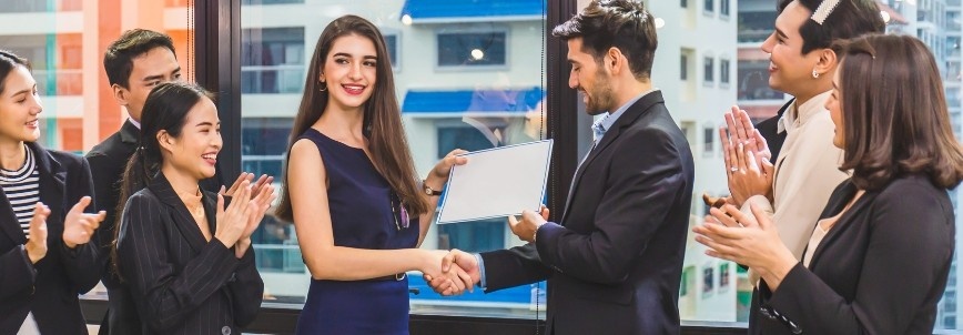 A woman shakes hands with a man while receiving a certificate, as colleagues applaud her CAPM® Certification achievement in a bright office setting with large windows and city buildings in the background.
