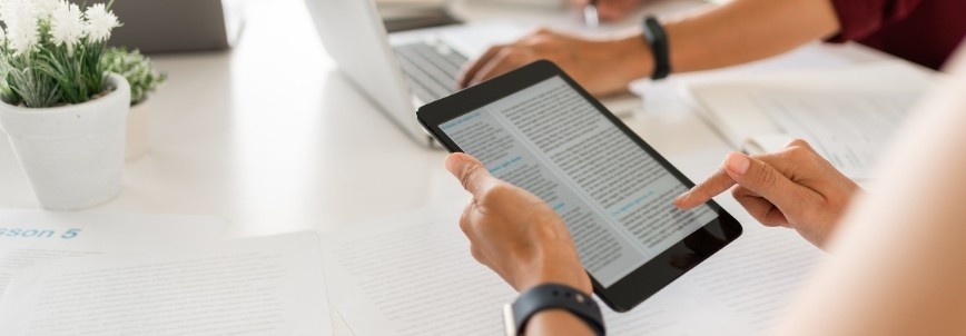 Two people work at a desk with documents and a laptop. One person scrolls through PMP Exam Questions on a tablet, while a small potted plant sits on the table beside the papers.