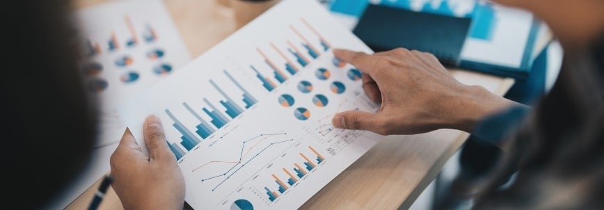 A person points to charts and graphs on a printed sheet, analyzing financial or business case data at a desk with documents and a notebook in the background.