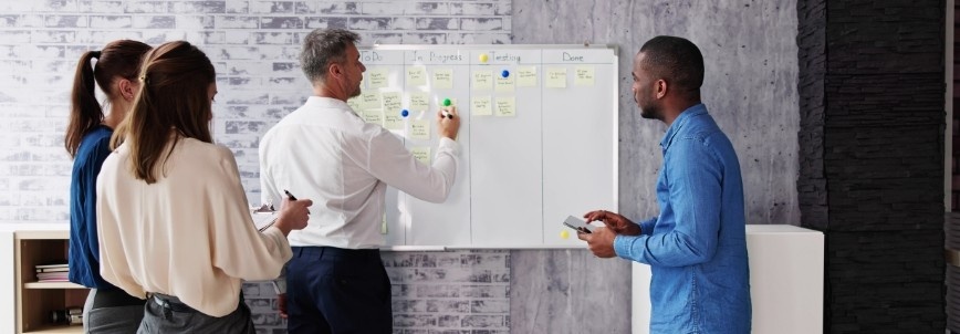 Four people stand in front of a whiteboard covered with sticky notes, collaborating on various tasks. As they discuss ideas and write notes, they explore different types of Agile projects in a modern office setting.