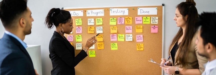 Four people stand by a corkboard with sticky notes in columns labeled "To Do," "In Progress," "Testing," and "Done." A Project Manager in Agile gestures toward the board while others listen attentively.