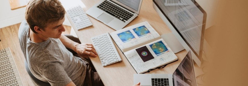 A person sits at a desk working on a laptop and desktop computer, surrounded by papers, keyboards, and a notebook displaying colorful charts—perhaps analyzing the ups and downs of online PMP® exams.
